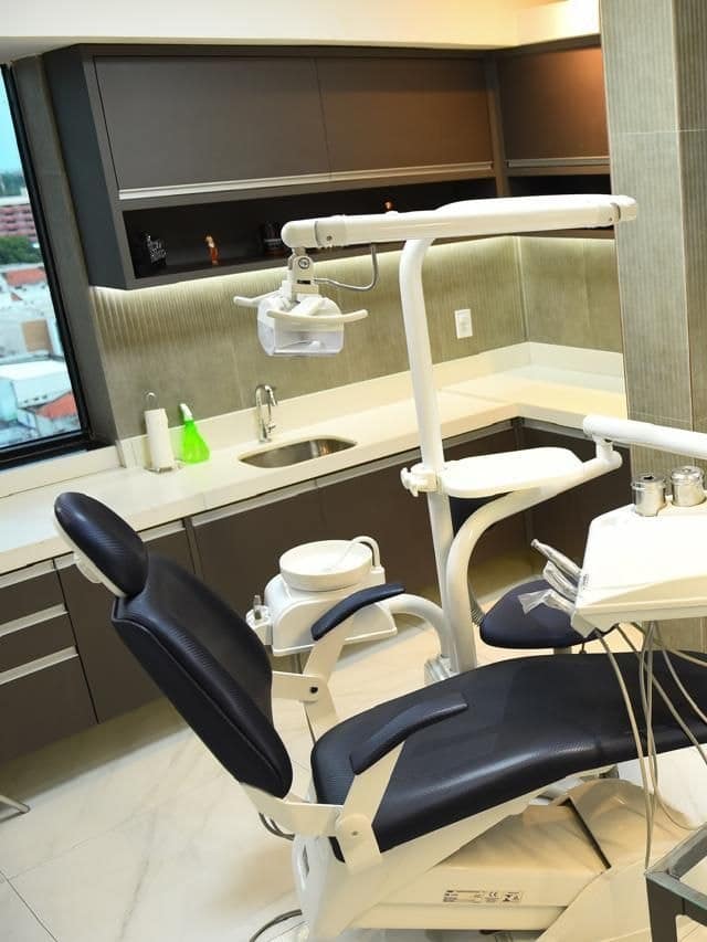 Sala clínica com cadeira odontológica, refletor, bancada e armários, iluminada por uma janela que dá para a cidade.